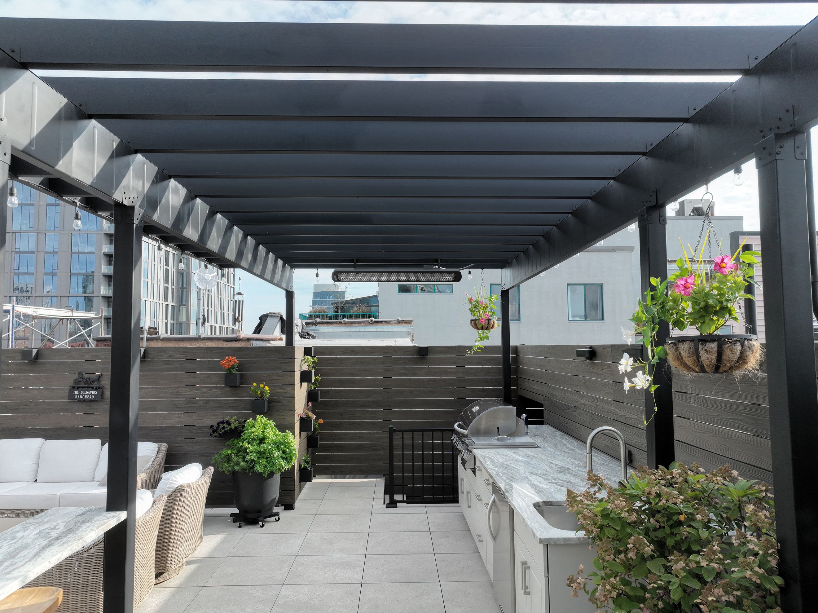 Outdoor steel pergola over outdoor kitchen space with buildings in the background.